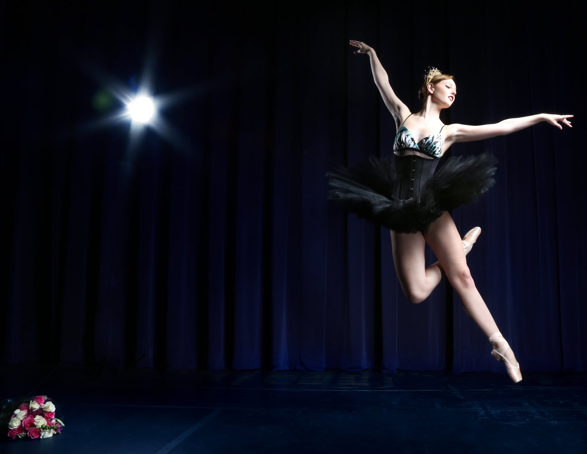 Ballerina jumping in mid-air against a dark curtain with a spotlight. She wears a black pancake tutu skirt, black corset, and teal silk bralette