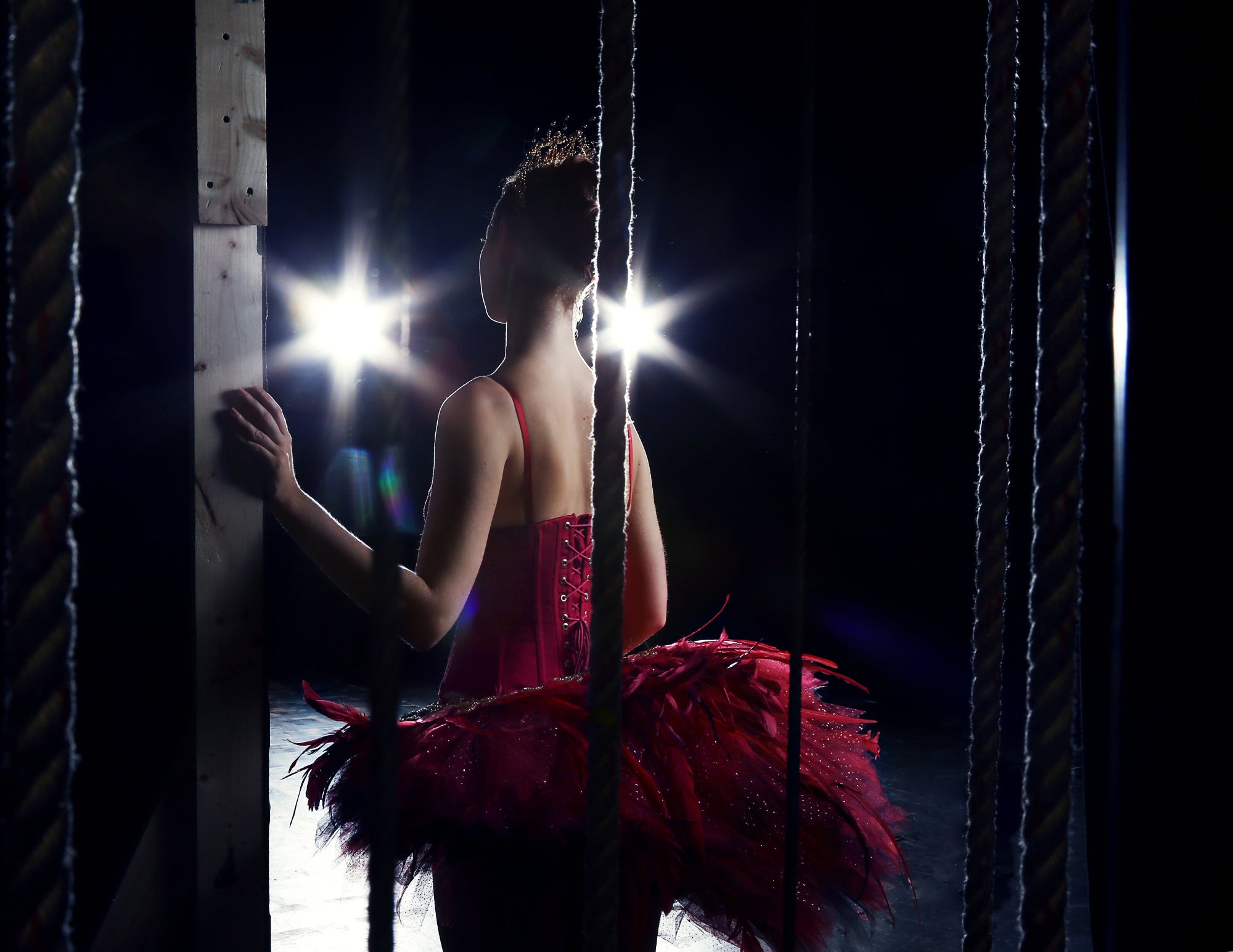 Ballerina in a red tutu standing behind ropes on a dark stage with spotlights.