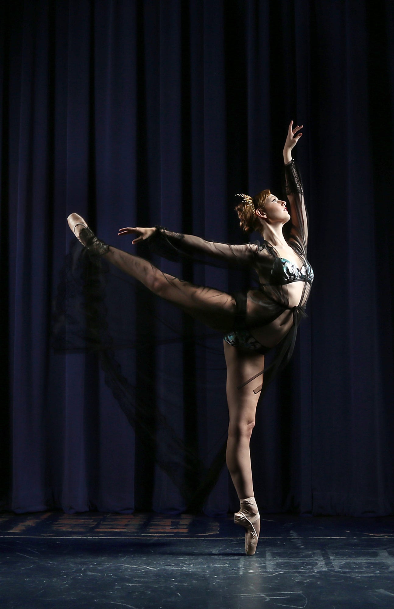 Ballerina posing in a high arabesque, wearing a black sheer tulle dress on stage