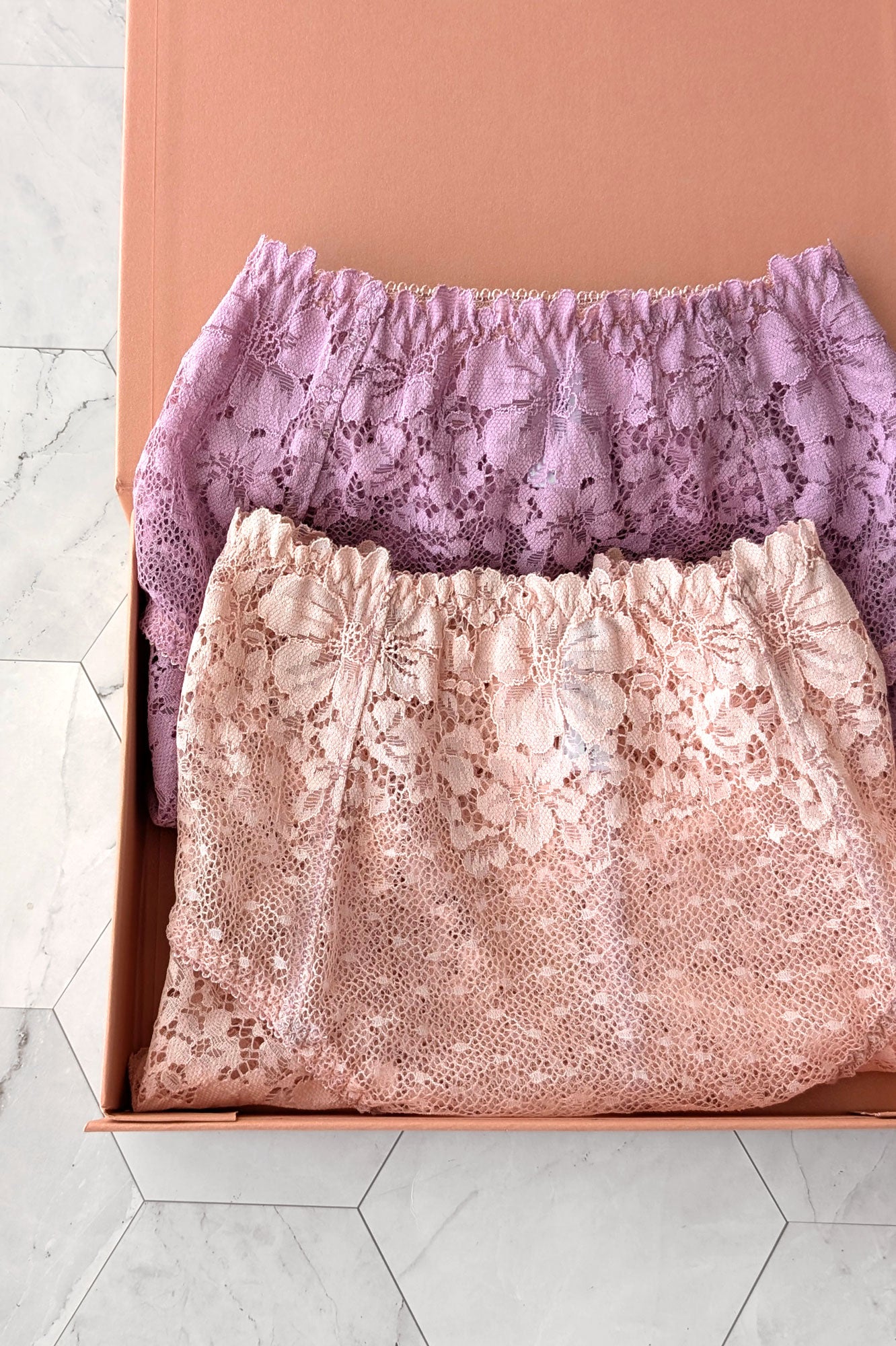 Two pairs of folded lace underwear, one light purple and one light pink, in a gift box on a marble floor.
