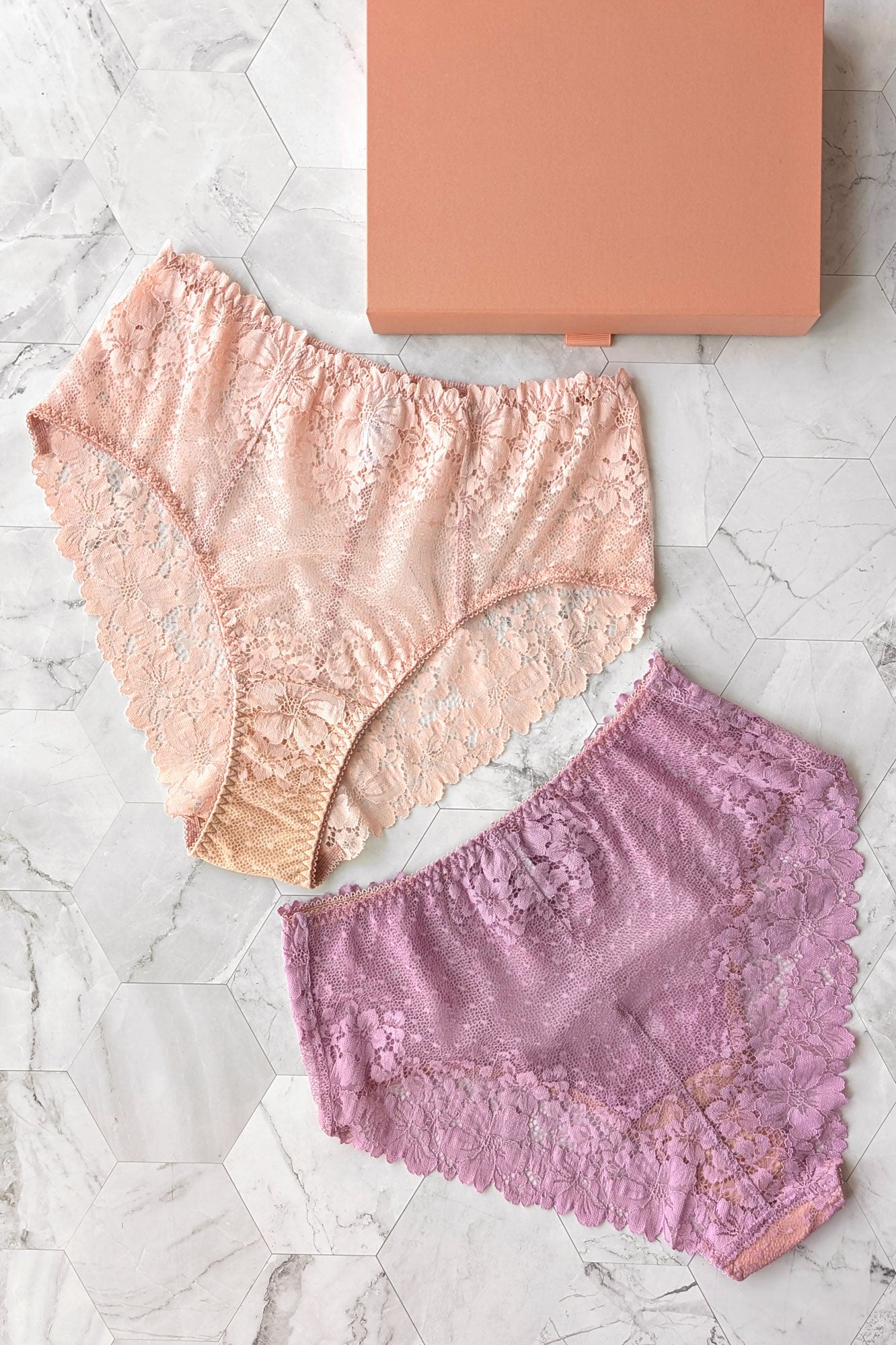 Two pairs of lace underwear, one pink and one purple, on a marble surface with a rose pink gift box.