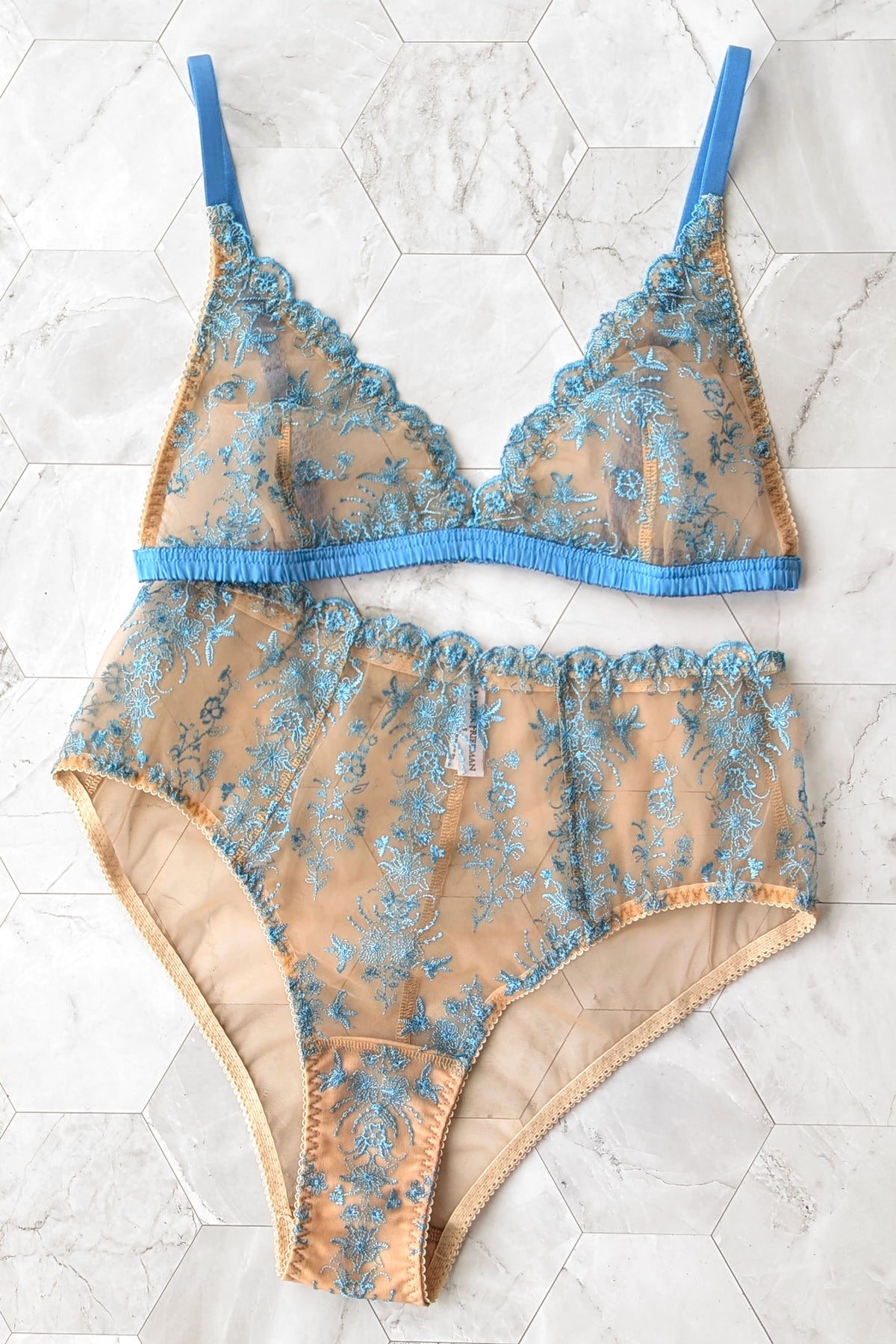 A two-piece sheer lingerie set featuring a bralette and matching high-waisted briefs in beige mesh with blue floral embroidery and azure blue silk trim