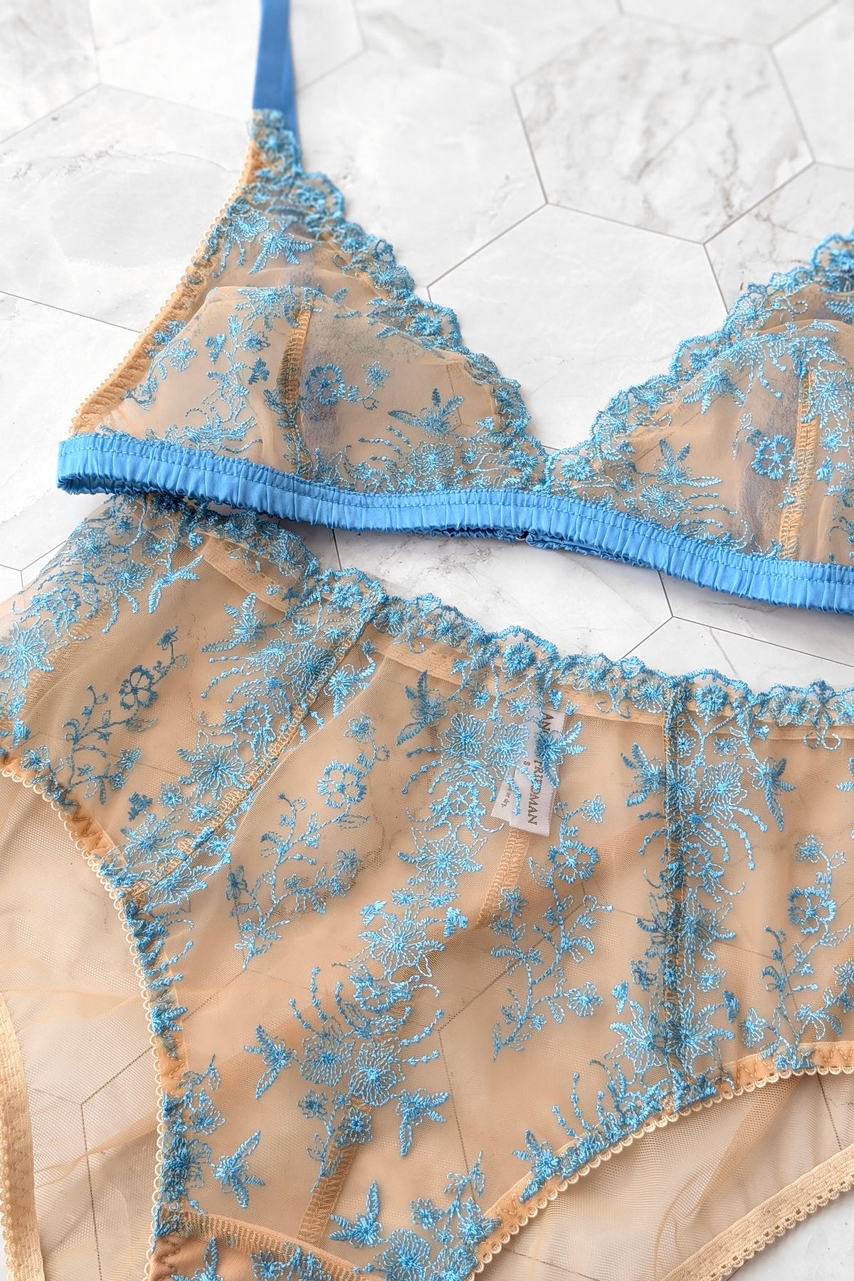 Close up photo showing the details of a 2-piece sheer lingerie set featuring a bra and matching panties in beige mesh with intricate light blue floral embroidery, laid flat on a light gray marble surface.