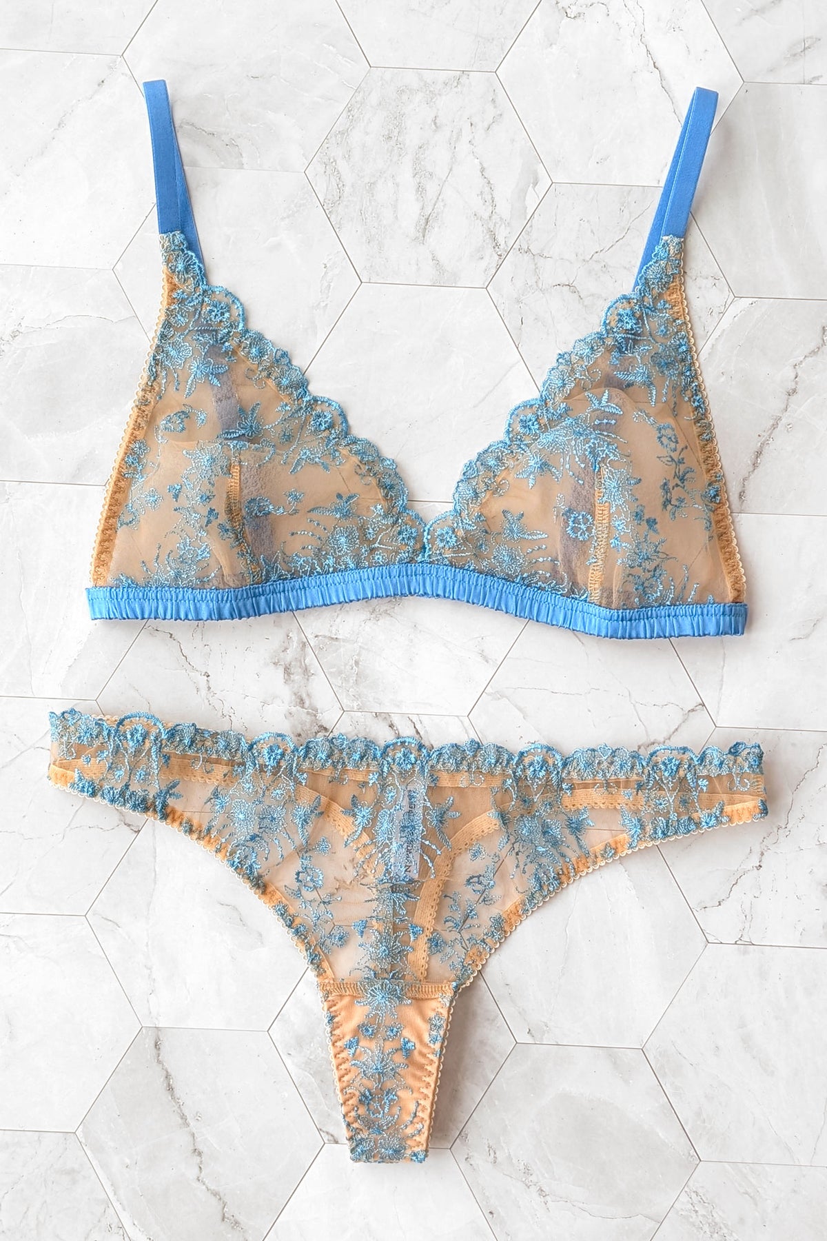 A two-piece sheer lace lingerie set featuring a triangle bra and matching thong in a peach-beige mesh with blue floral embroidery and solid blue straps, laid flat on a white hexagonal tile background