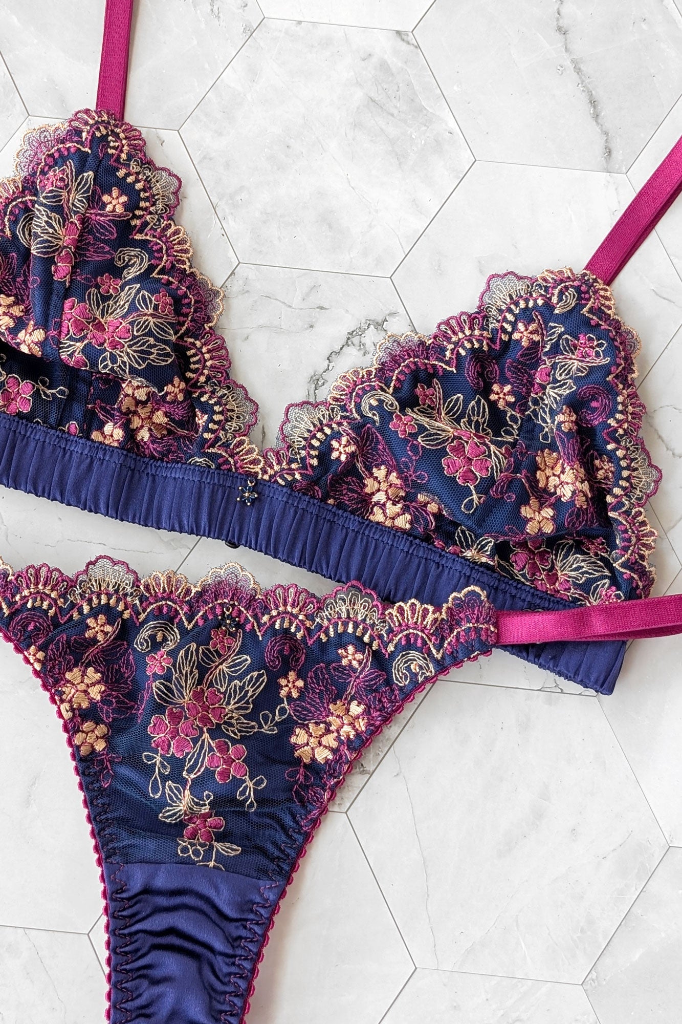 Floral embroidered lingerie set in blue silk with bright pink elastic straps on a white marble background