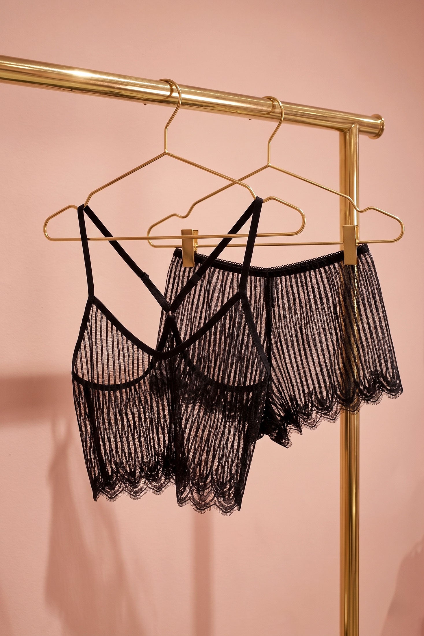 Luxury designer lingerie set in sheer black French lace, hanging on gold hangers against a pink background