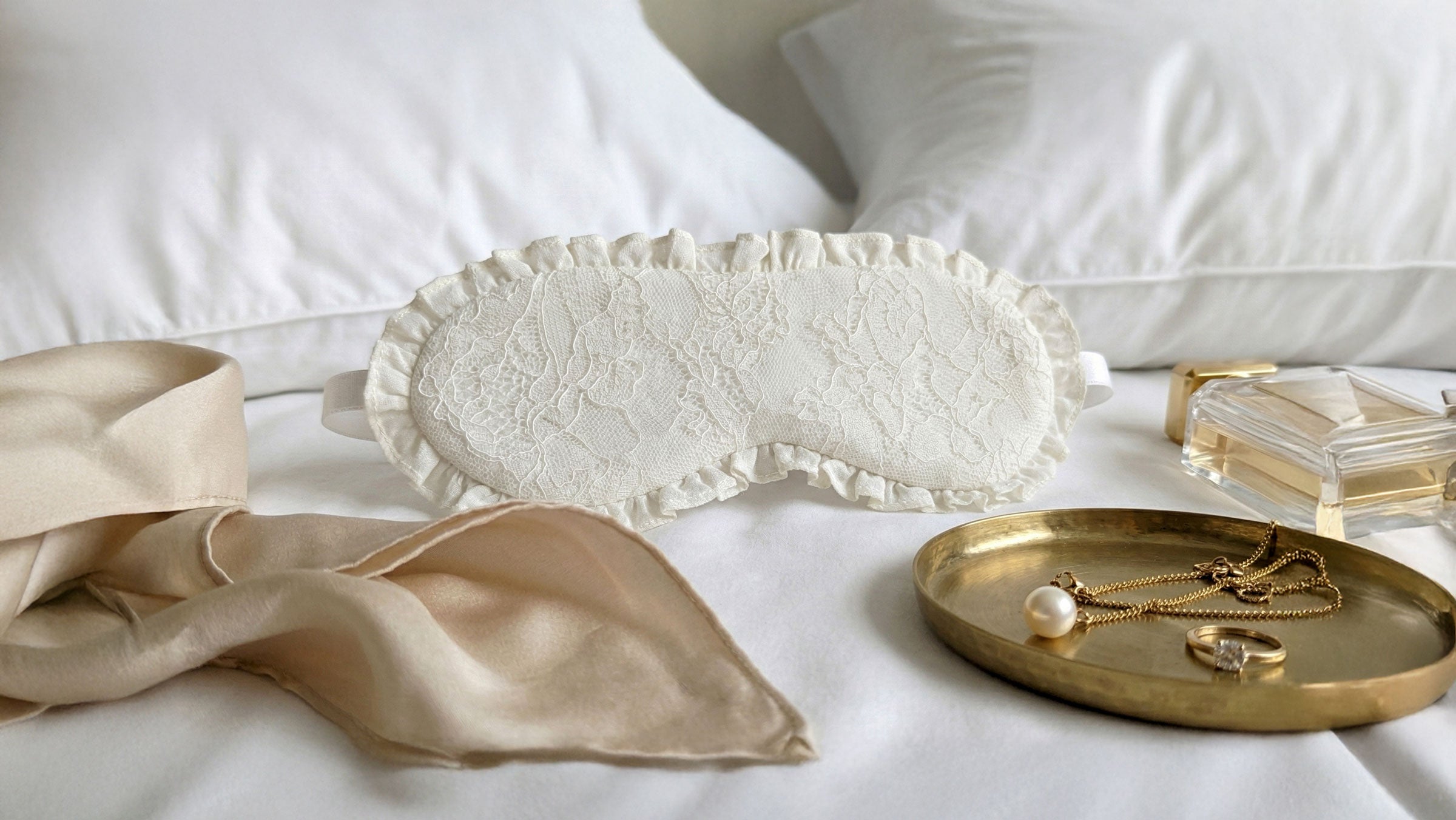 white lace sleep mask for brides on her wedding day, laid on a white hotel bed with gold jewellery and perfume