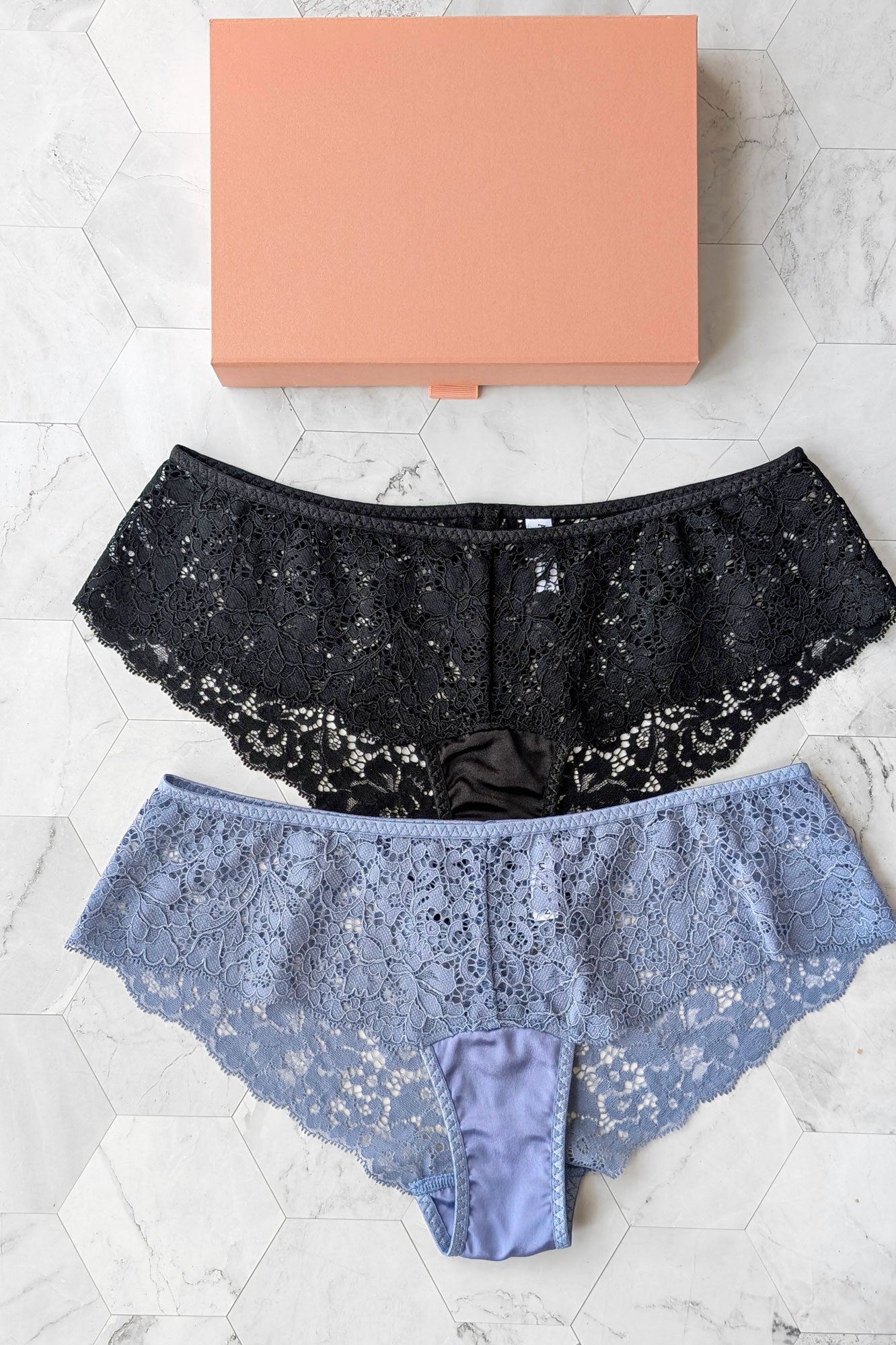 Two pairs of silk and lace underwear, one jet black and one thistle blue, on a white marble surface with a peach-colored gift box in the background.