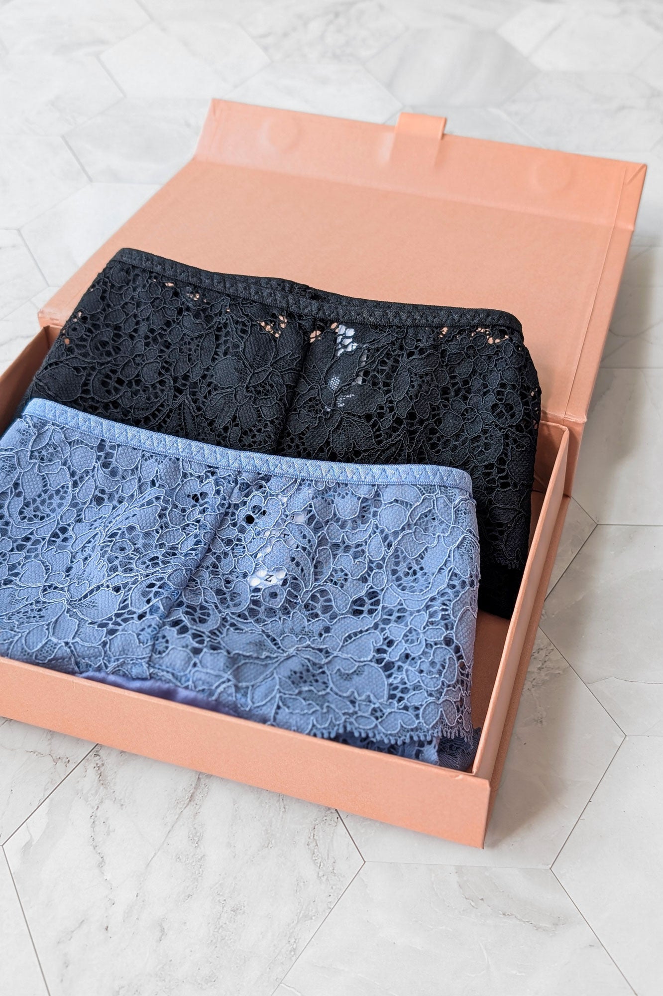Two folded pairs of lace panties, one black and one blue, in a rose pink luxury magnetic gift box, ready for gift giving.
