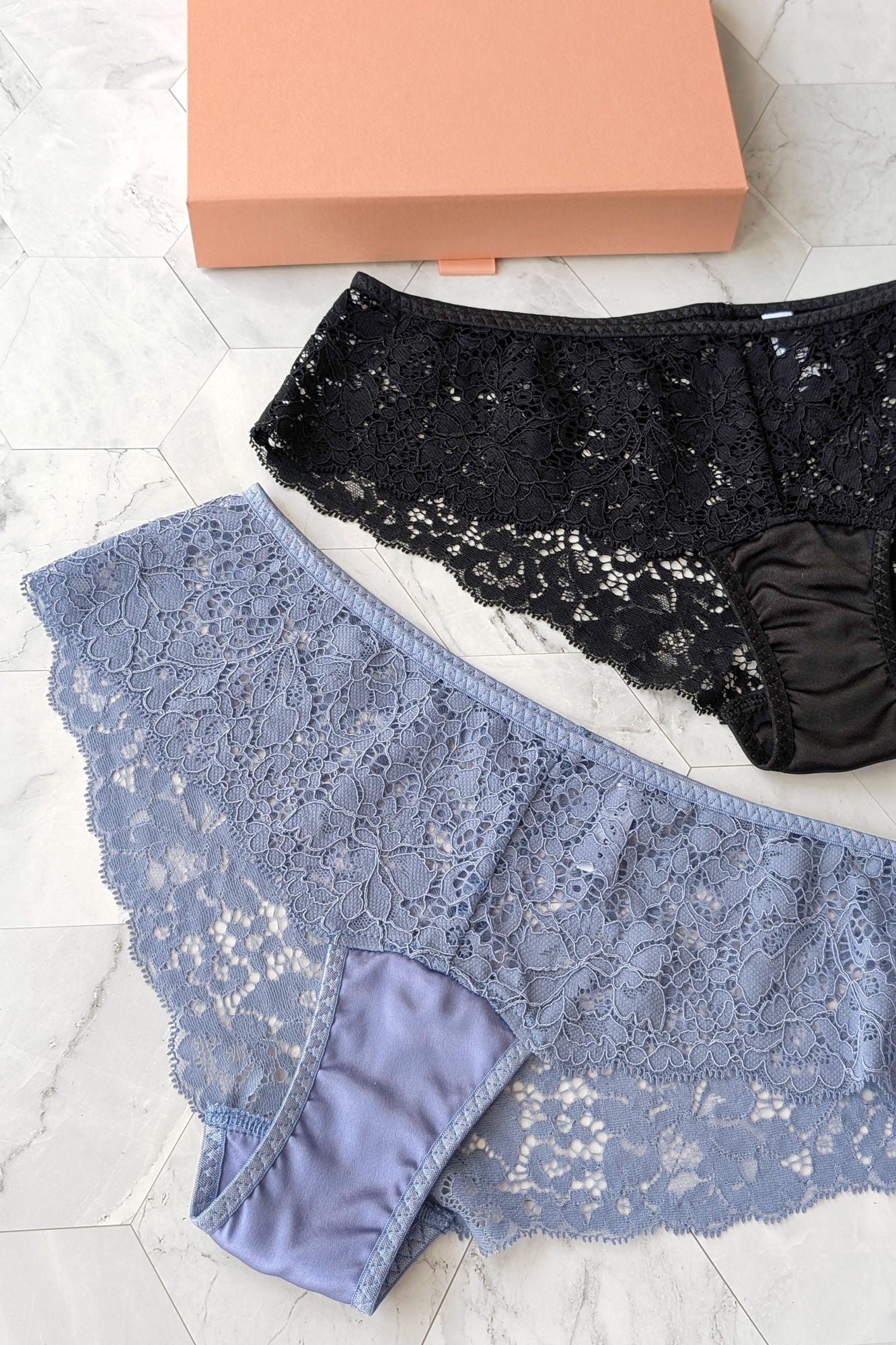 Flat lay image of black lace underwear and gray blue lace underwear on a white surface with a pink box in the background.