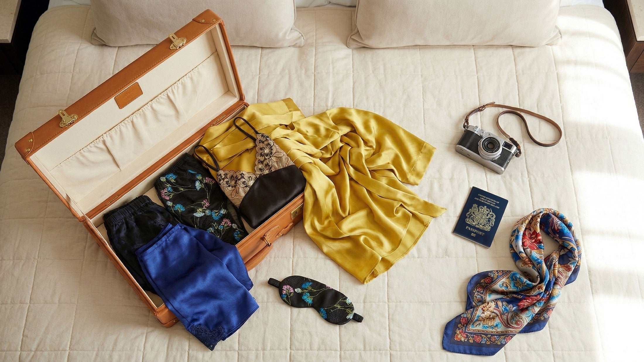 100% silk robes and loungewear pieces spilling out of a vintage suitcase on a hotel bed