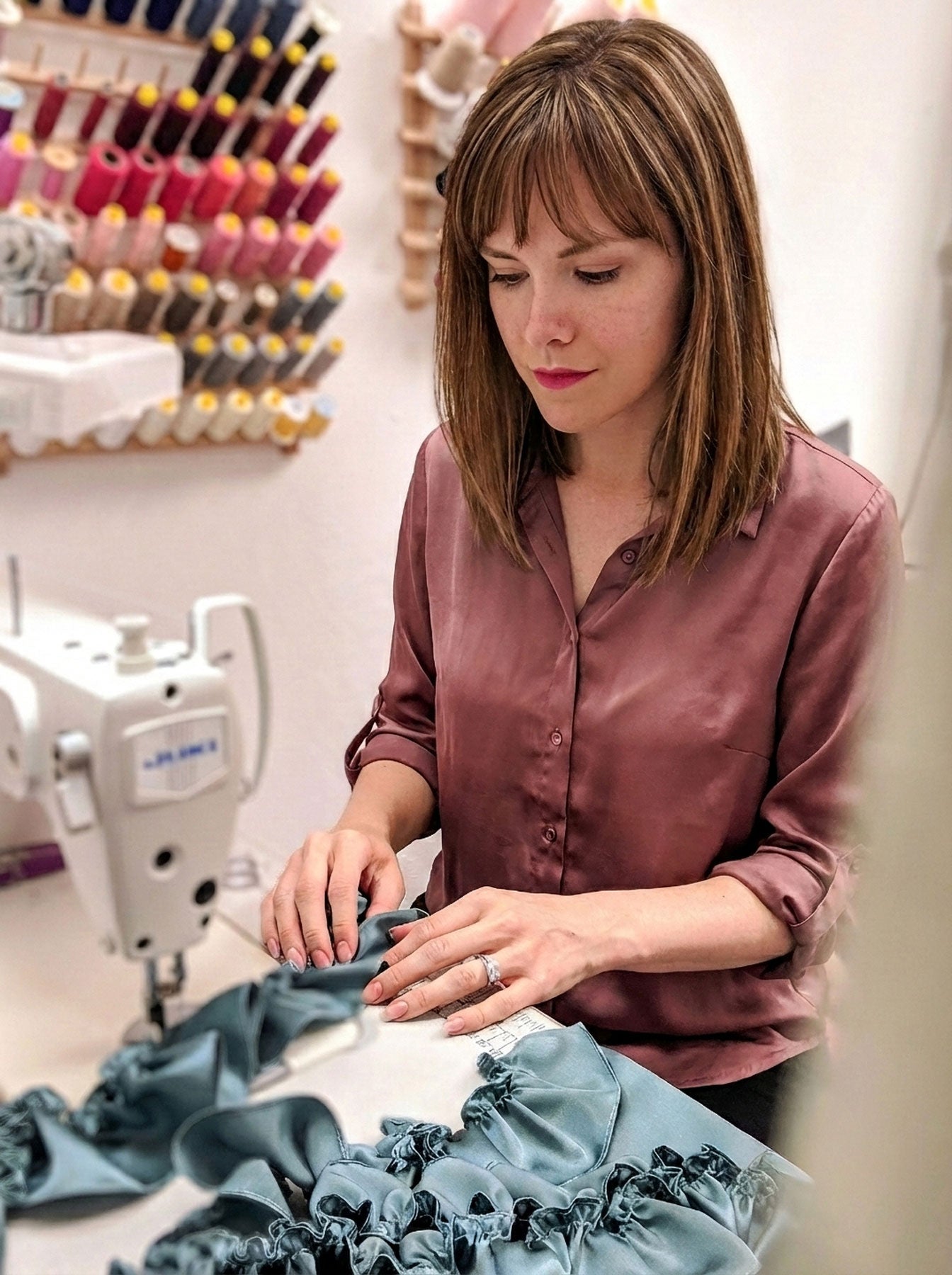 Lingerie Designer Angela Friedman working on a sewing project in a workshop with sewing machine and thread spools in the background.