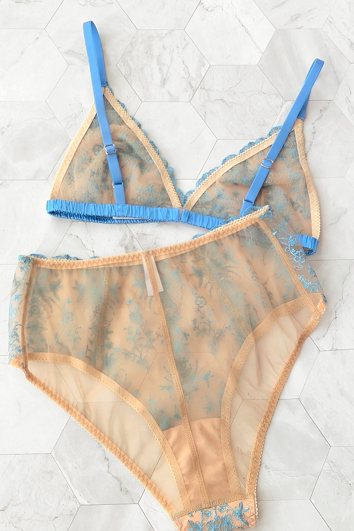 A two-piece sheer lingerie set laid flat on a light marble tile background. The set includes a bralette and matching high-waisted briefs in a peach-beige color mesh with a light blue floral pattern and solid blue elastic straps