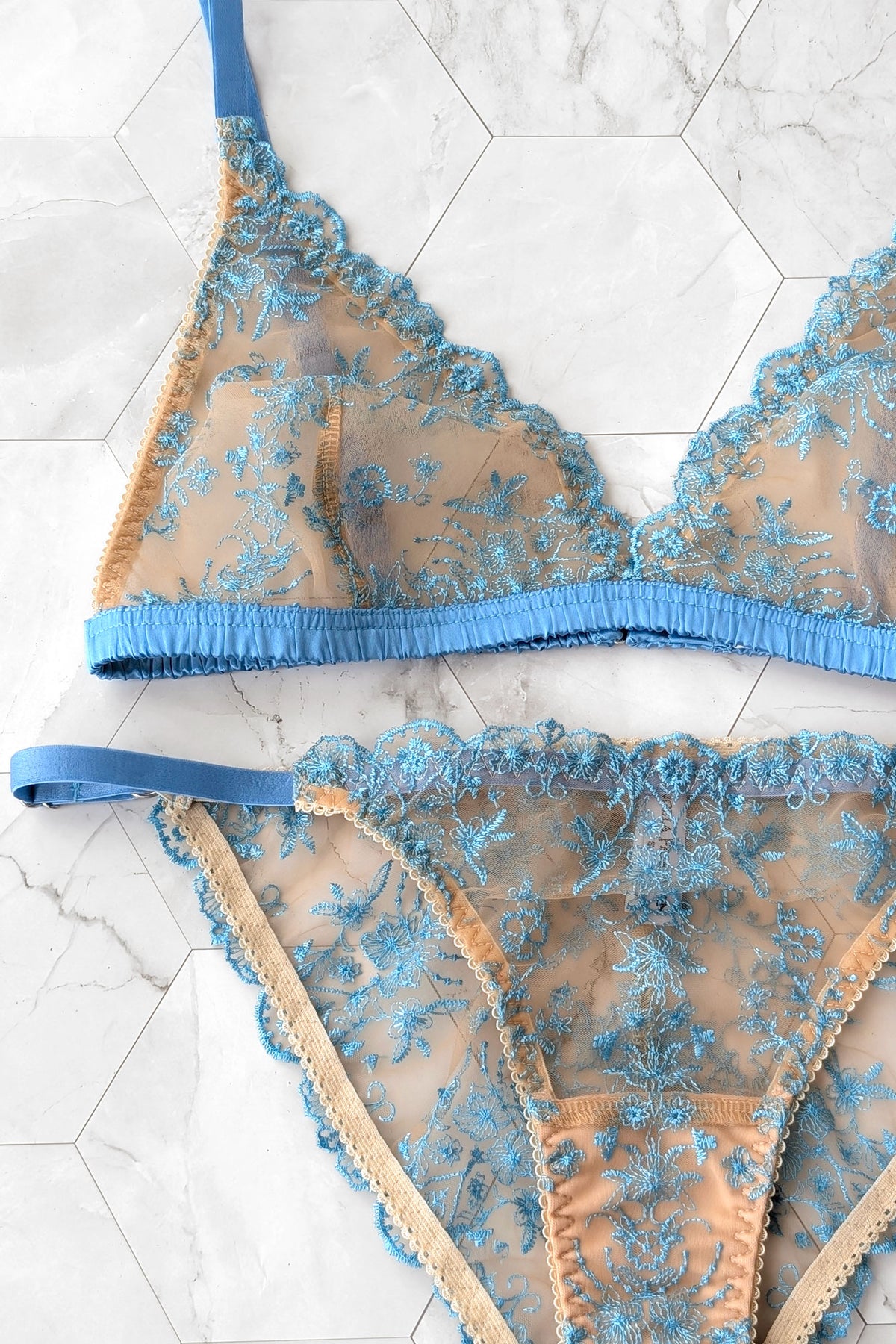 Close up photo of a 2-piece lace lingerie set featuring a sheer mesh bralette and matching high-waisted briefs with intricate blue floral embroidery, blue 100% silk trim, and blue straps