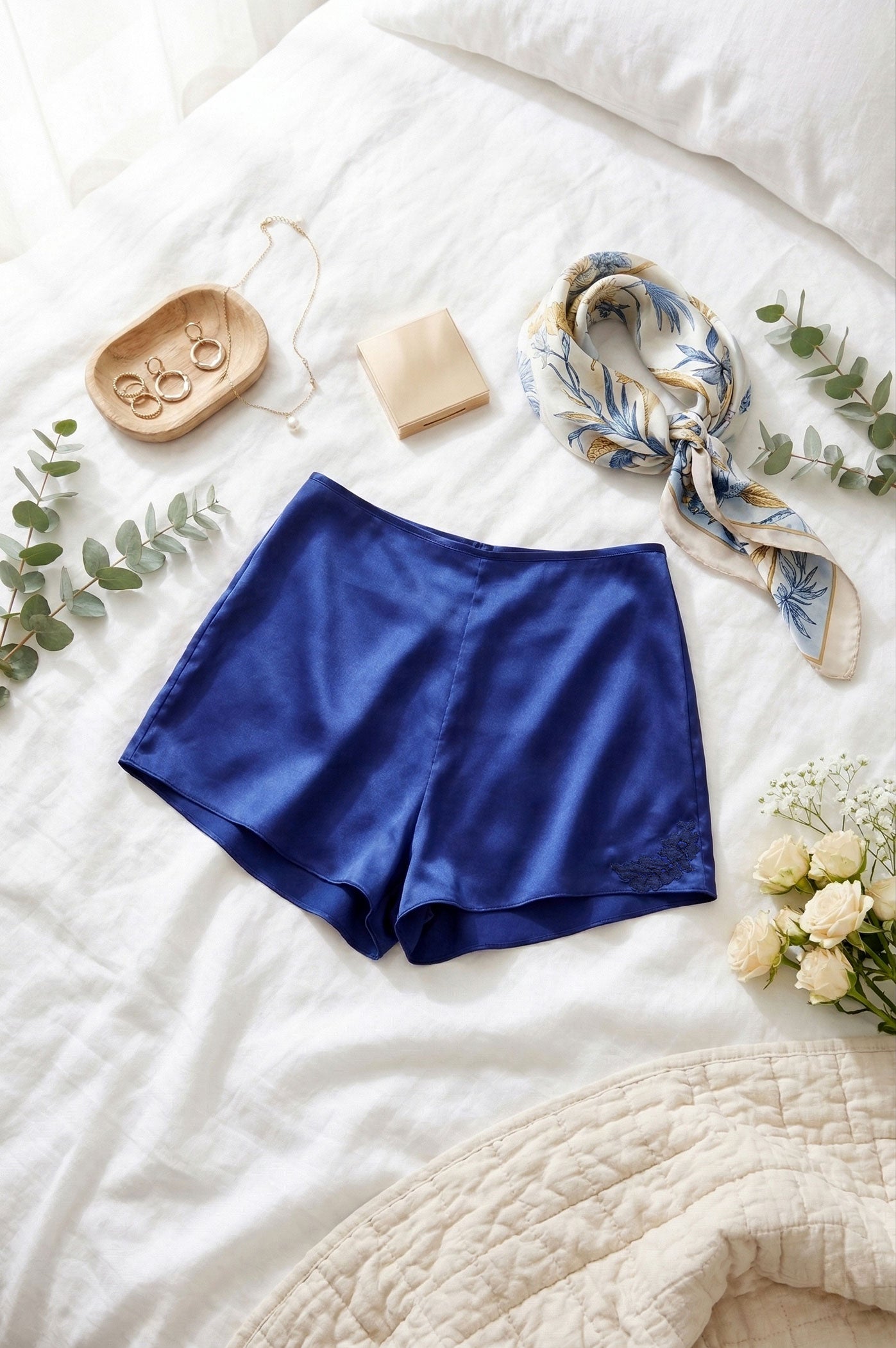 Blue satin sleep shorts on a white bed with a printed silk scarf, jewelry, and flowers