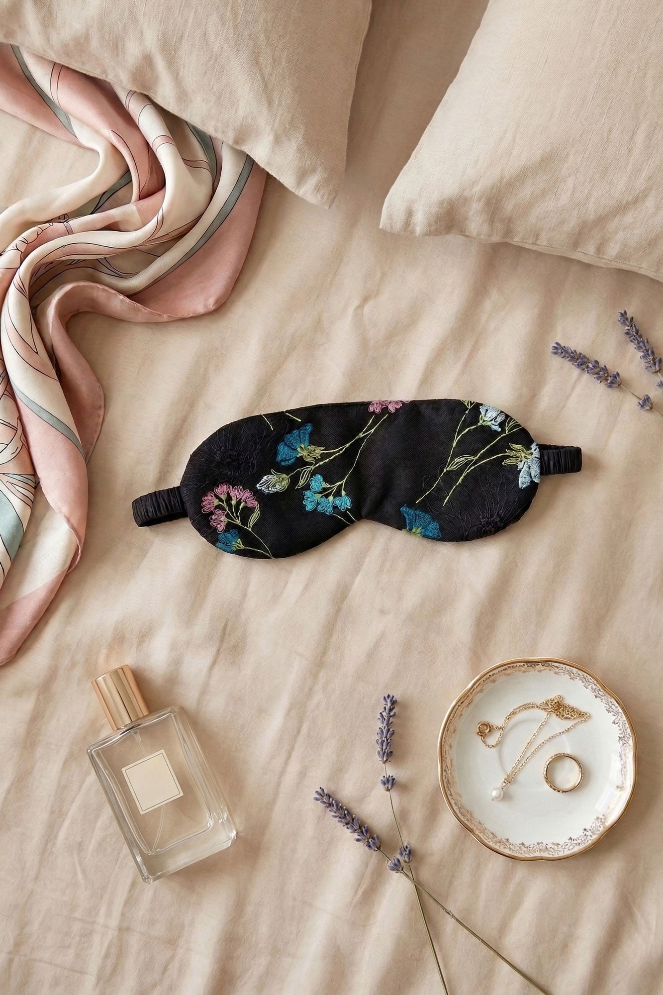Black sleep mask with floral embroidery on a beige linen bed with bedtime accessories