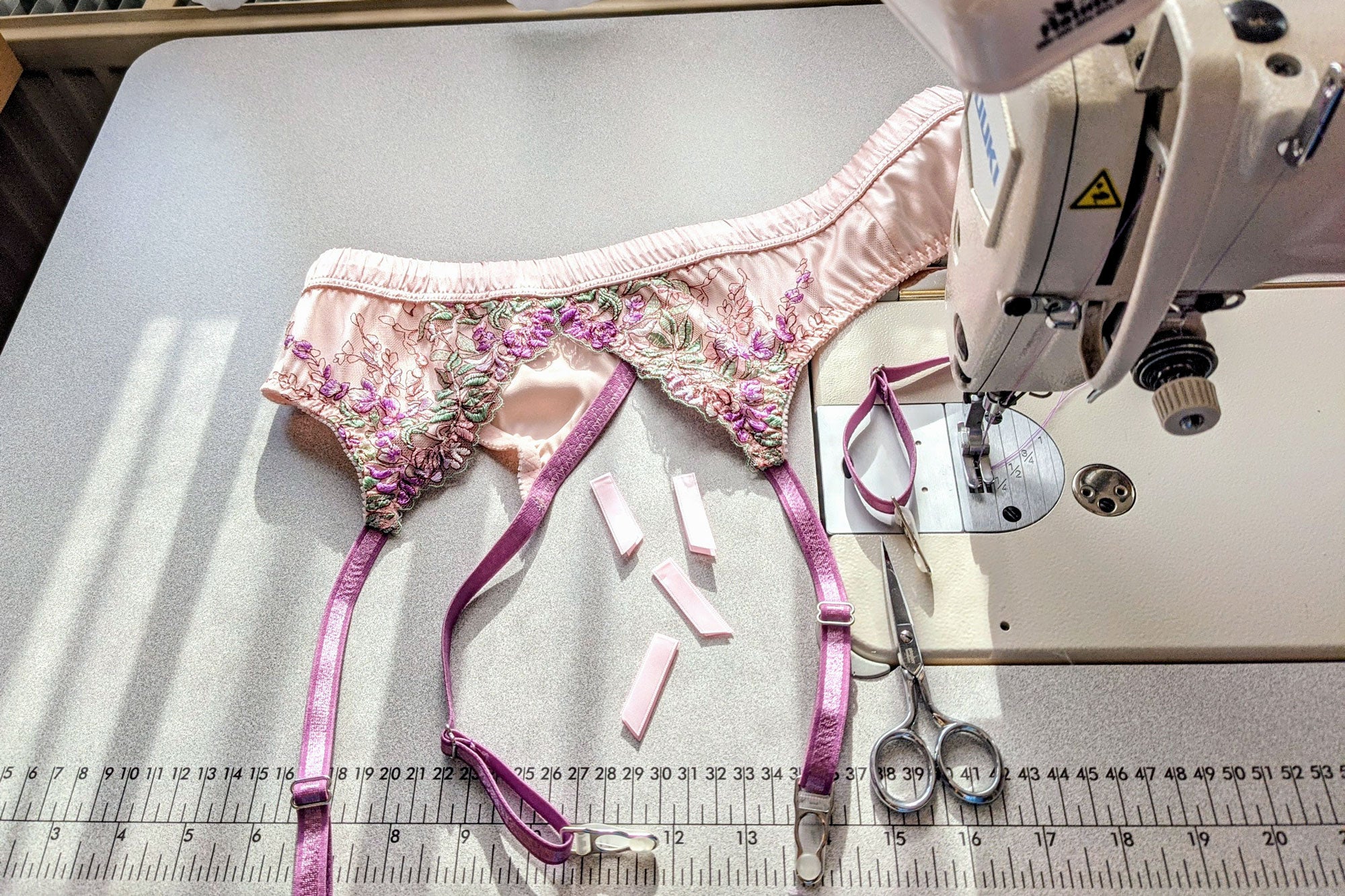 Pink garter belt on a sewing machine table, unfinished with scissors and suspender ribbon tabs remaining to be sewn