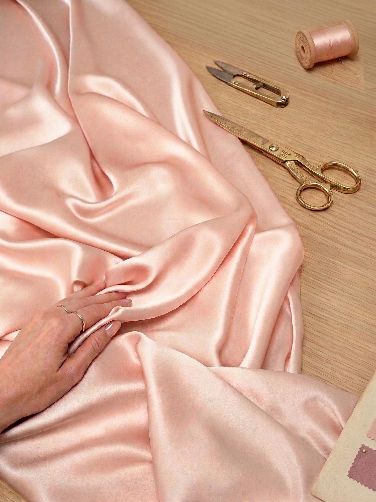 Pink silk satin fabric with scissors and a spool of thread on a wooden surface