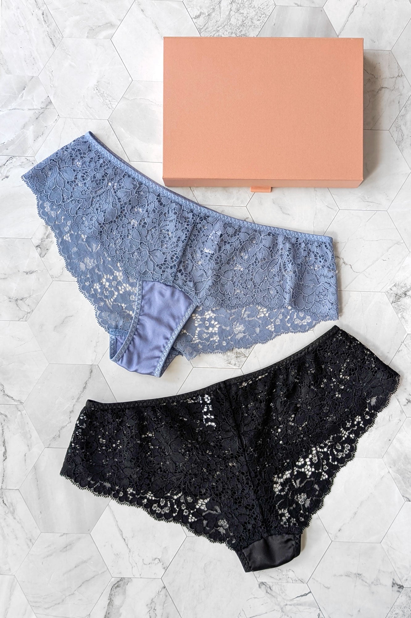 Two pairs of lace underwear, one blue and one black, on a textured white surface with a rose-gold-colored box.